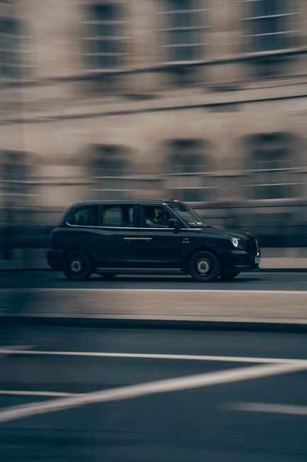 A black London-style taxi cab is moving along a city street with a background of tall, modern buildings appearing blurred due to the motion. The vehicle is in the midst of a home relocation process, p